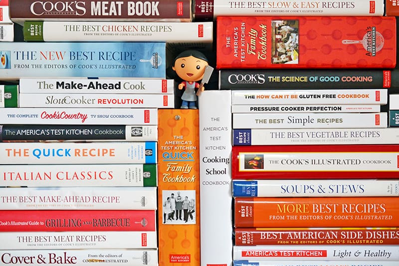 A big stack of Cook's Illustrated and America's Test Kitchen cookbooks with a Mini Michelle action figure in the middle.