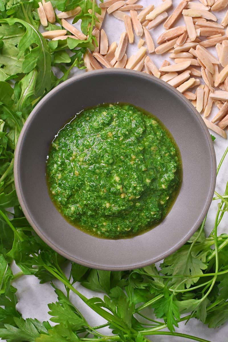 A small bowl containing arugula pesto with the ingredients of arugula and slivered almonds surrounding the bowl.