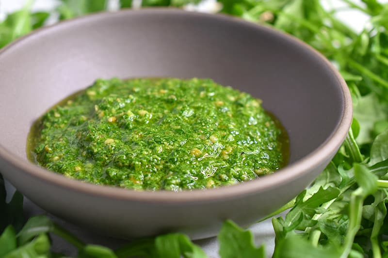 A small bowl filled with arugula pesto with arugula surrounding the bowl.