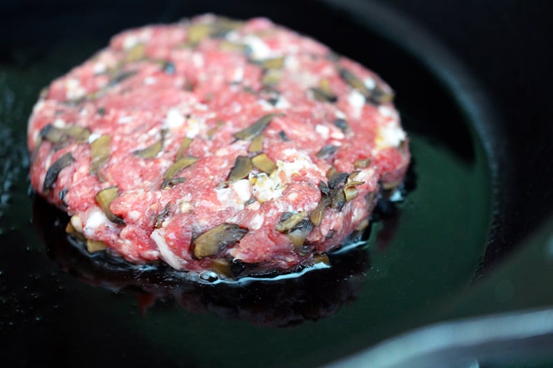 A patty of big-o bacon burger frying in a cast iron skillet.