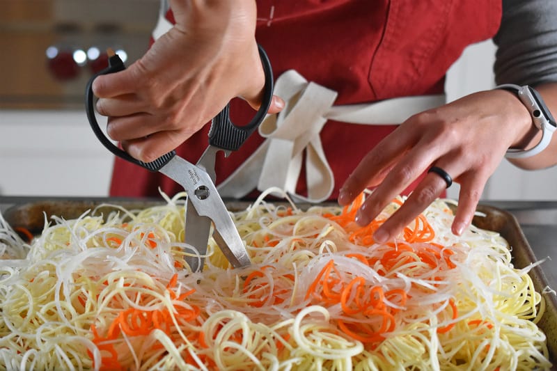 Spiralized white sweet potatoes, carrots, and onions are on a rimmed baking sheet and they are being cut with a pair of kitchen shears.