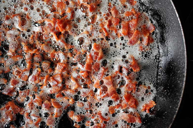 An overhead shot of bacon bits frying in a large cast iron skillet.