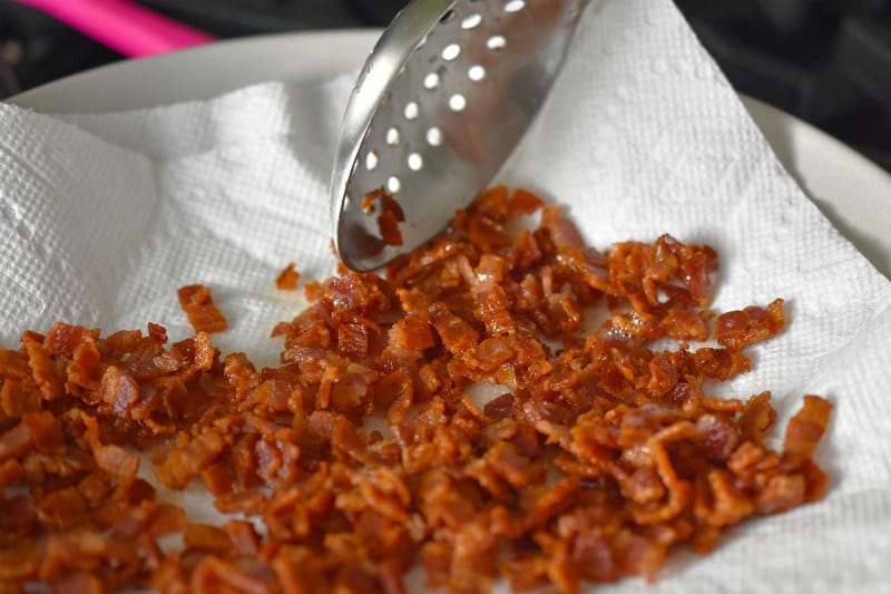 Crispy bacon bits are transferred to a paper towel-lined plate with a slotted spoon.