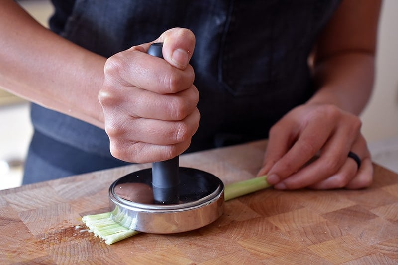 Smashing a piece of trimmed lemongrass with a metal meat pounder on a wooden cutting board.