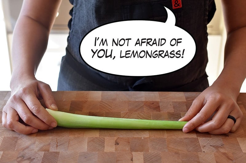 A person holding a stalk of lemongrass on a wooden cutting board with a cartoon word bubble that says, "I’m not afraid of you, lemongrass!"