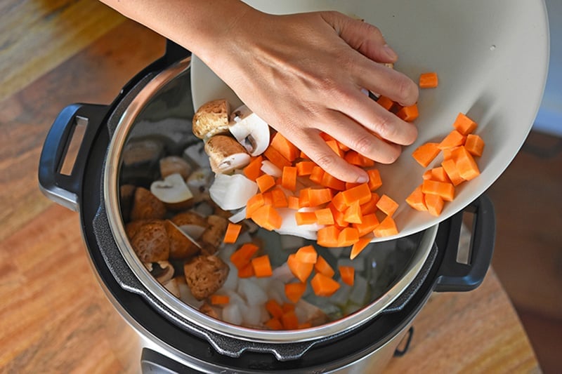 An overhead shot of someone adding mushrooms, onions, and carrots to an open Instant Pot.