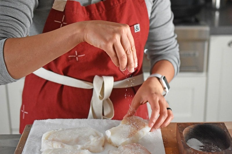 A woman in a red apron is sprinkling kosher salt on raw cod fillets.