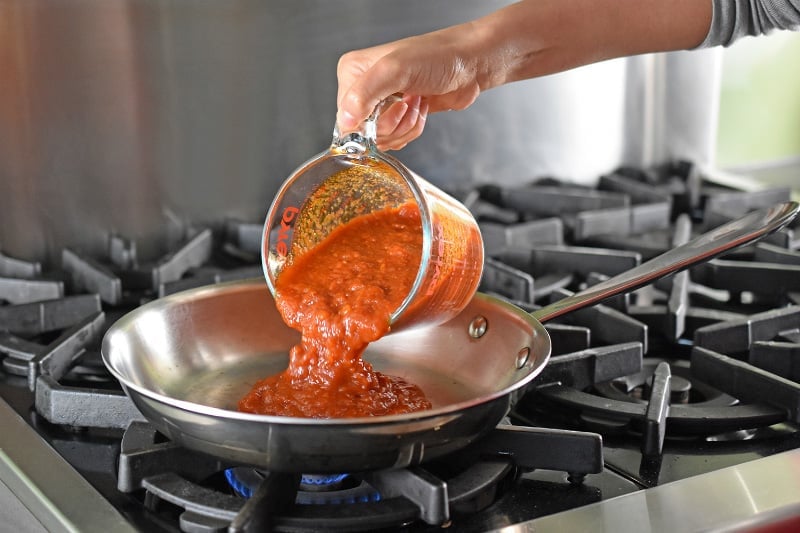Marinara sauce is poured from a Pyrex measuring cup into a large stainless steel skillet on a stovetop.