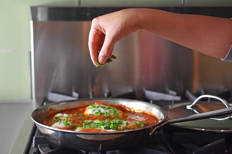 Sprinkling fresh herbs on a skillet filled with Poached Cod in Tomato Sauce 