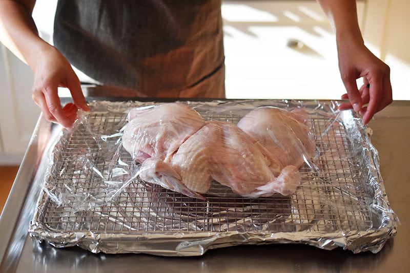 A raw spatchcocked chicken on a wire rack in a rimmed baking sheet is covered with plastic wrap