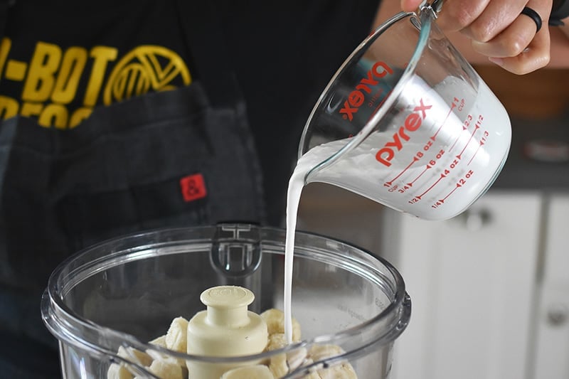 Someone pouring coconut milk into a food processor with frozen banana pieces.