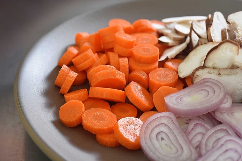 A platter with sliced carrot coins, sliced shiitake mushrooms, and sliced shallots.