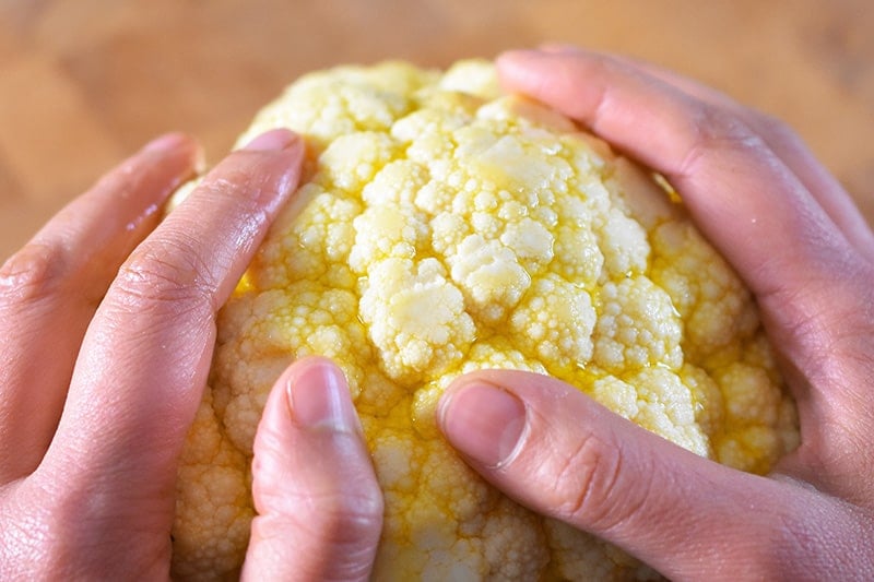 Two hands are massaging olive oil onto the surface of a small head of cauliflower.
