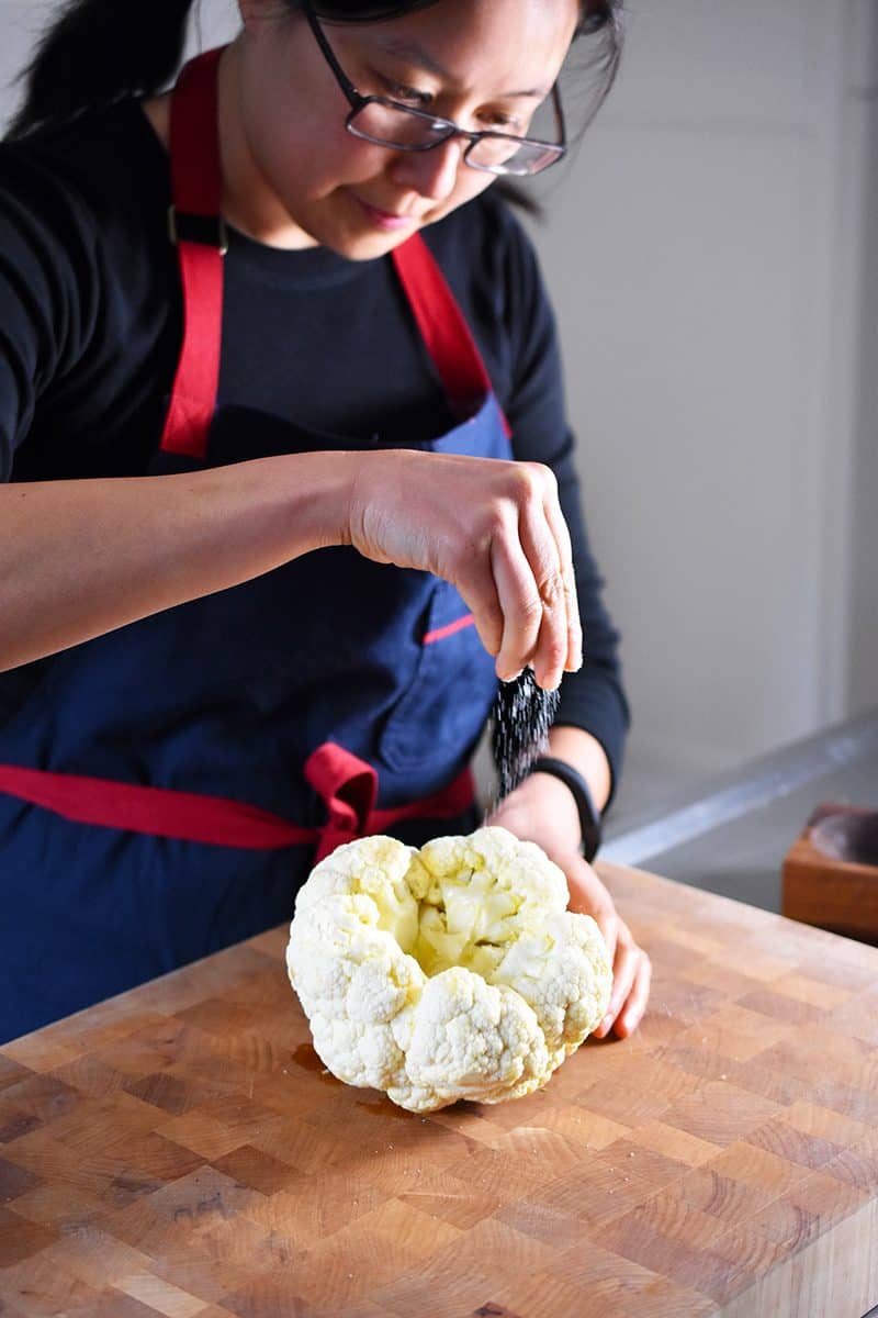 A woman is sprinkling salt all over a whole cauliflower head coated with olive oil.