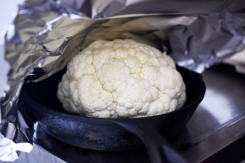 The seasoned cauliflower head is in a small cast iron skillet and aluminum foil is placed on top.