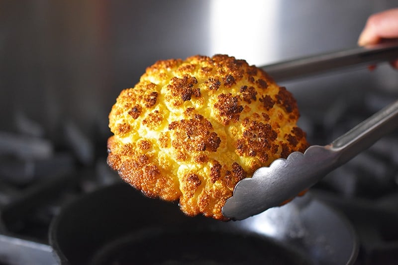 A pair of tongs remove Whole Roasted Cauliflower from the cast iron skillet.