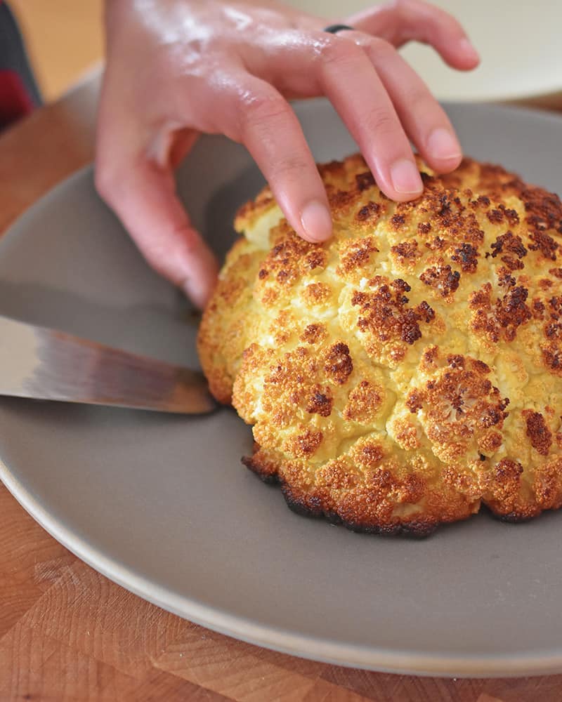 A shot of someone slicing Whole Roasted Cauliflower on a gray plate.