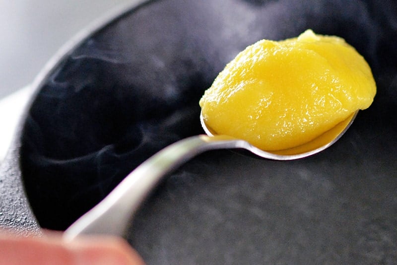 A spoon filled with ghee is on top of a smoking hot cast iron skillet.