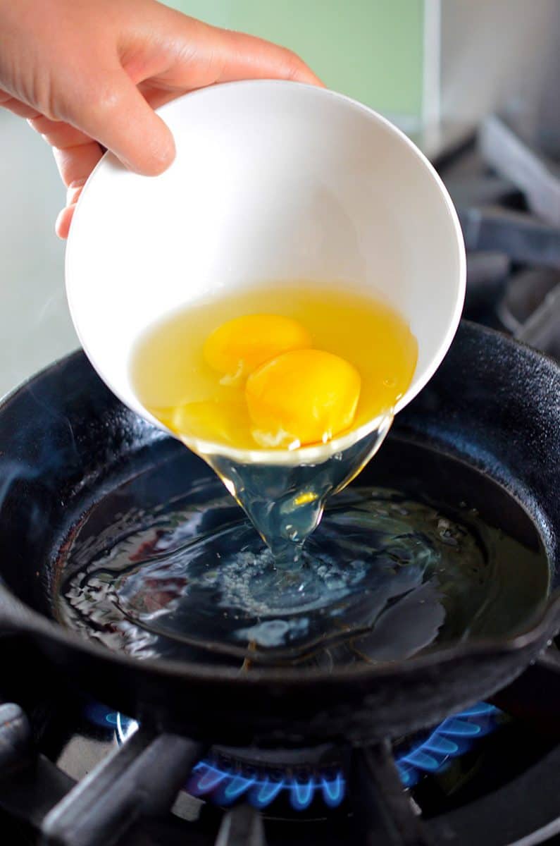 Two eggs in a white bowl are poured into a hot cast iron skillet.