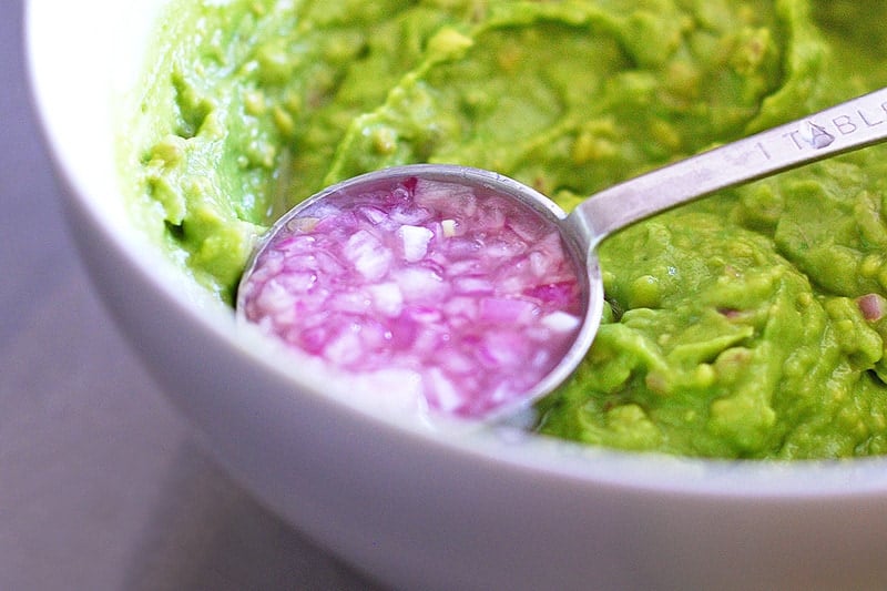 A tablespoon filled with the lime-shallot mixture is being added to the mashed avocado.