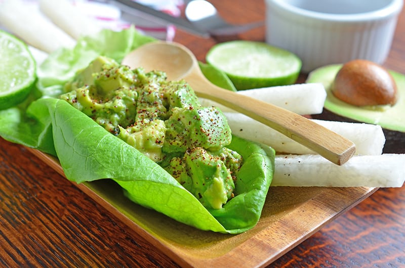 A lettuce taco is shown with guacamole inside, along with some jicama sticks, lime halves, and serving spoons.