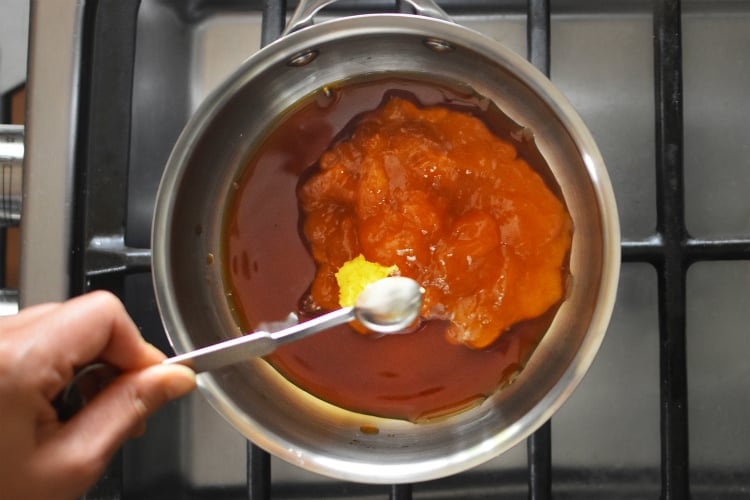 An overhead shot of someone adding a spoonful of minced ginger to a saucepan filled with ingredients for homemade Whole30 Apricot-Ginger Sauce 