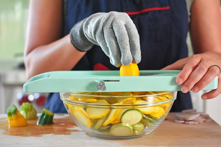 A gloved hand is using a green Japanese mandolin to thinly slice summer squash for Lemon Garlic Chicken Parchment Packets 