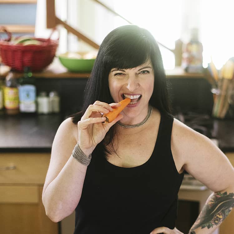 Picture of Melissa Joulwan, the author of Well Fed Weeknights, eating a carrot in her kitchen.