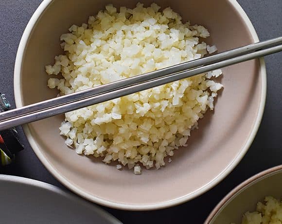 An overhead shot of cauliflower rice in a bowl topped with silver chopsticks