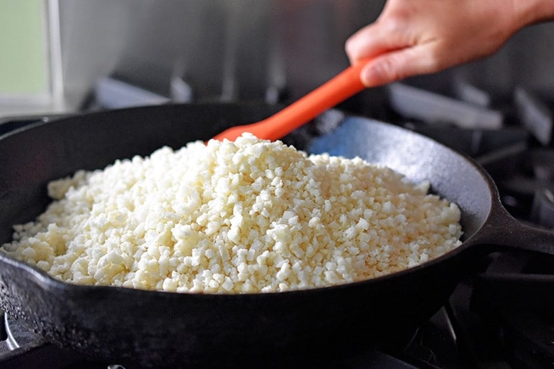A large cast iron skillet filled with riced cauliflower, being stirred with a red silicone spatula. 
