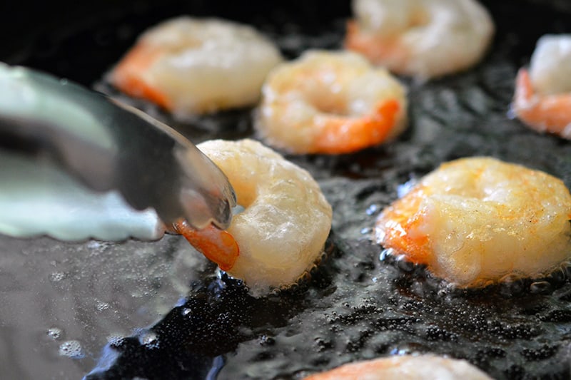 Using tongs to flip golden brown Paleo Walnut Shrimp frying in a cast iron skillet.