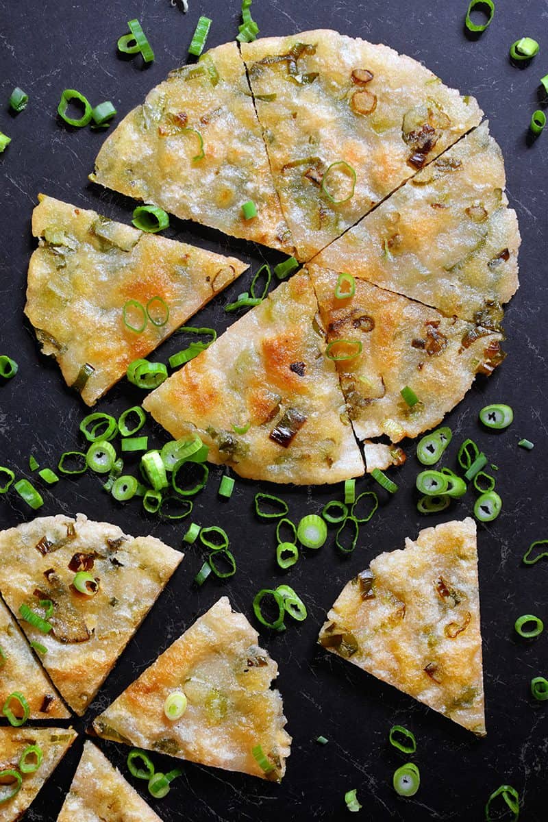 An overhead shot of Paleo Scallion Pancakes cut into wedges on a black background what sliced scallions scattered around them.
