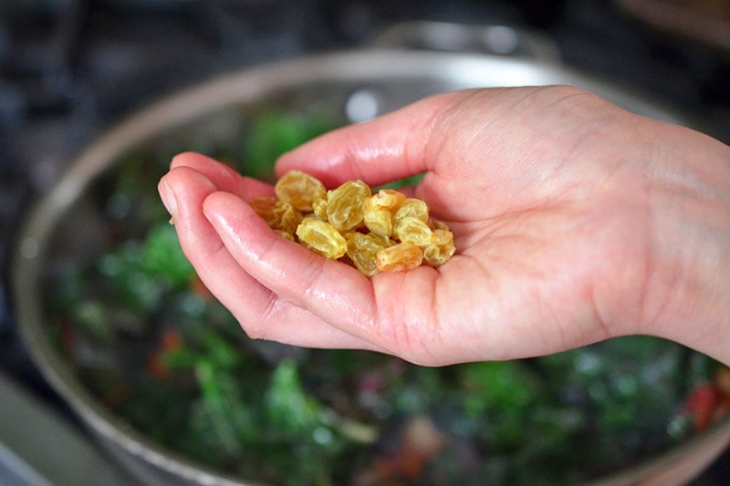 Someone holding a handful of plumped golden raisins to be added to a pan with sautéed swiss chard.