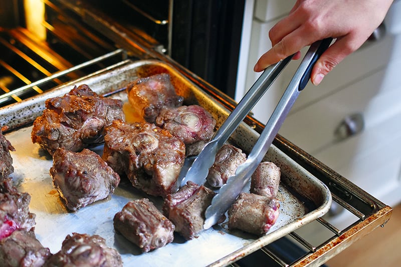 Broiled oxtails being flipped with a pair of tongs to brown evenly.