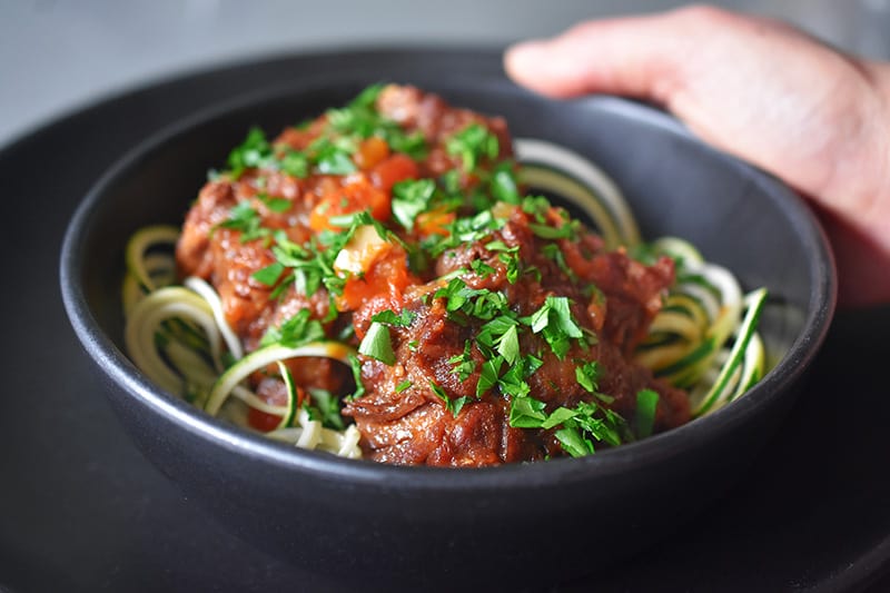A side view of someone holding a black bowl filled with zoodles topped with Instant Pot Oxtail Stew.