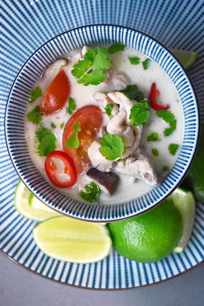 An overhead shot of a bowl of paleo and Whole30 Tom Kha Gai in a bowl on top of a plate with sliced limes.