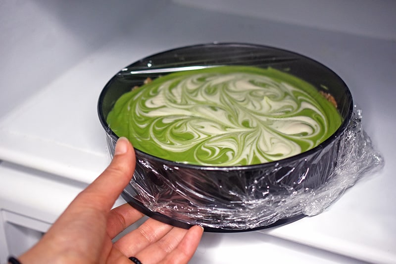 A paleo No-Bake Matcha Cheesecake covered with plastic wrap is placed in the refrigerator to chill.