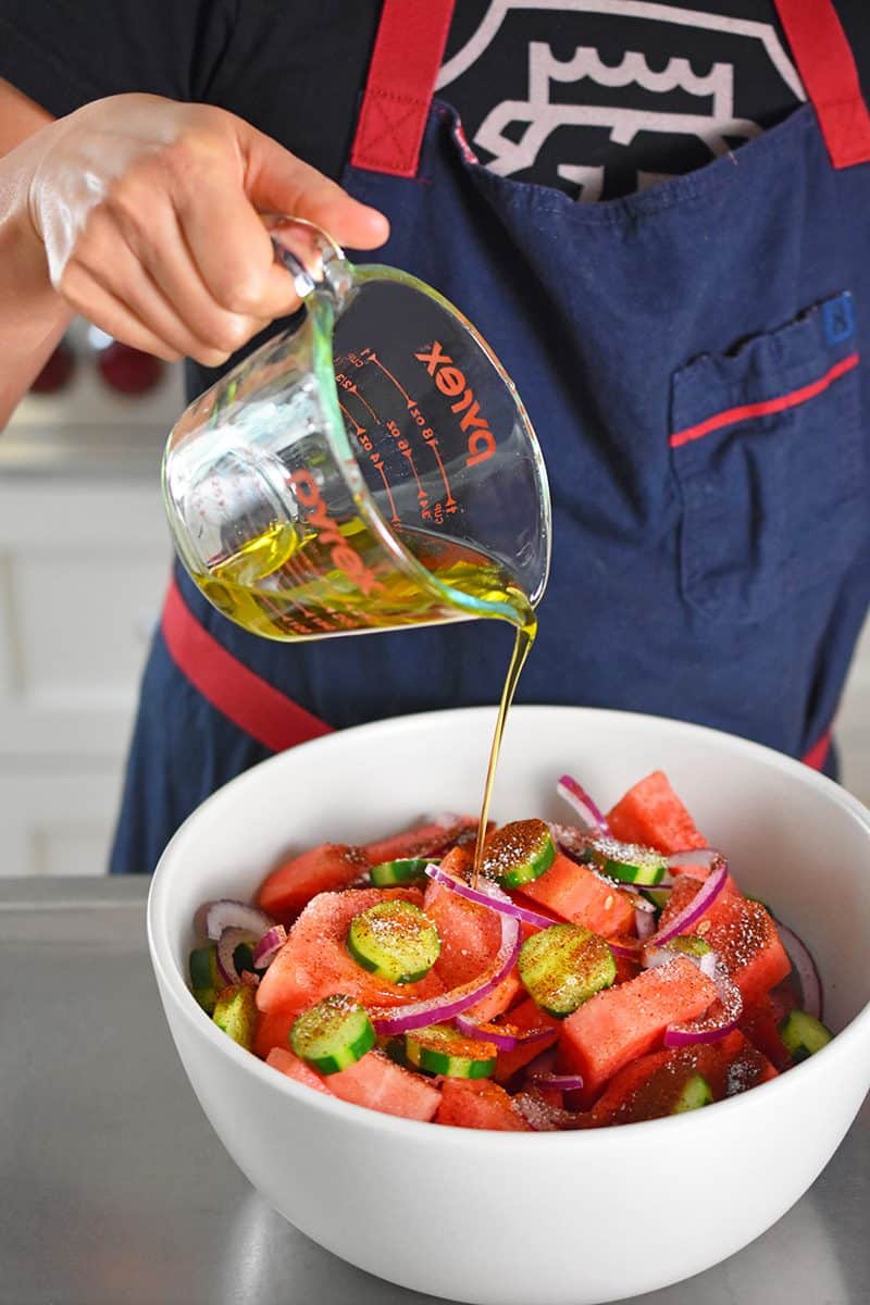 Someone pouring olive oil into a white bowl with the ingredients for the paleo Mexican watermelon salad.