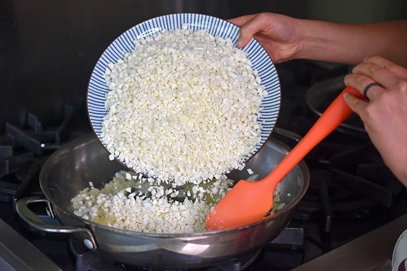 A person pouring a plate of riced cauliflower into a large stainless steel skillet.