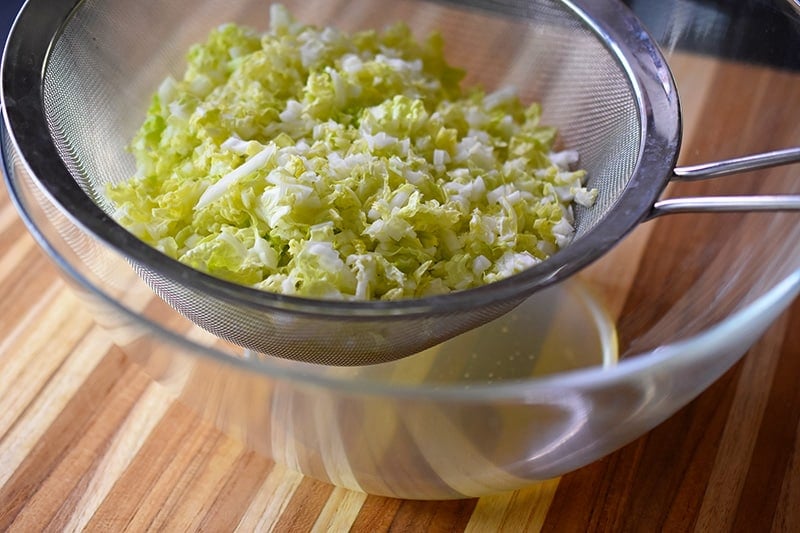 The salted napa cabbage in the fine mesh sieve releases liquid into the bowl.