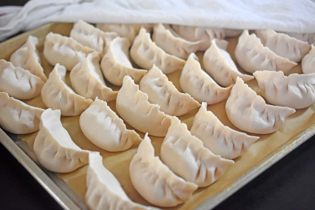A parchment-lined rimmed baking sheet is filled with Paleo Pot Stickers. There is a damp towel covering part of them.
