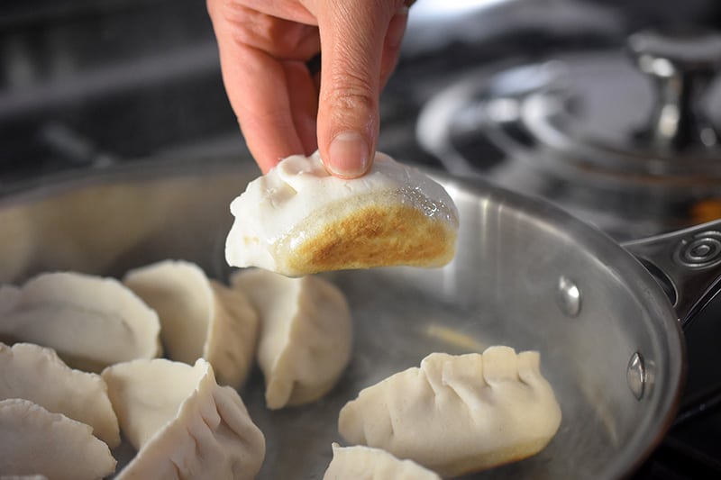 A hand is picking up a pot sticker from the skillet and the bottom of the dumpling is golden brown.