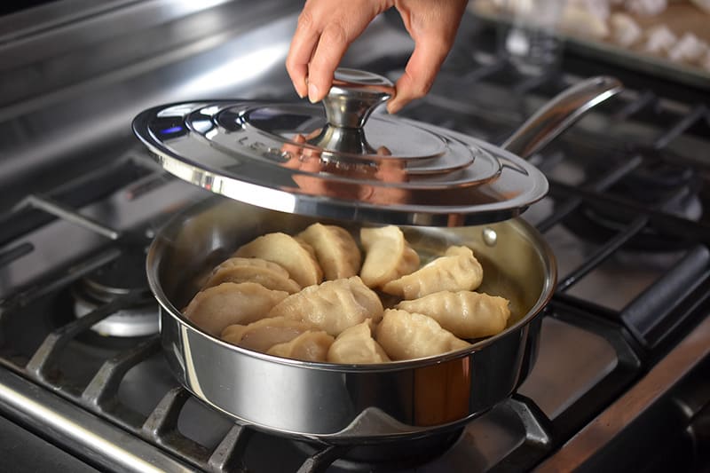 The lid being removed from a finished pan of Paleo Pot Stickers. The Pot Stickers are puffy and the skins are glossy.