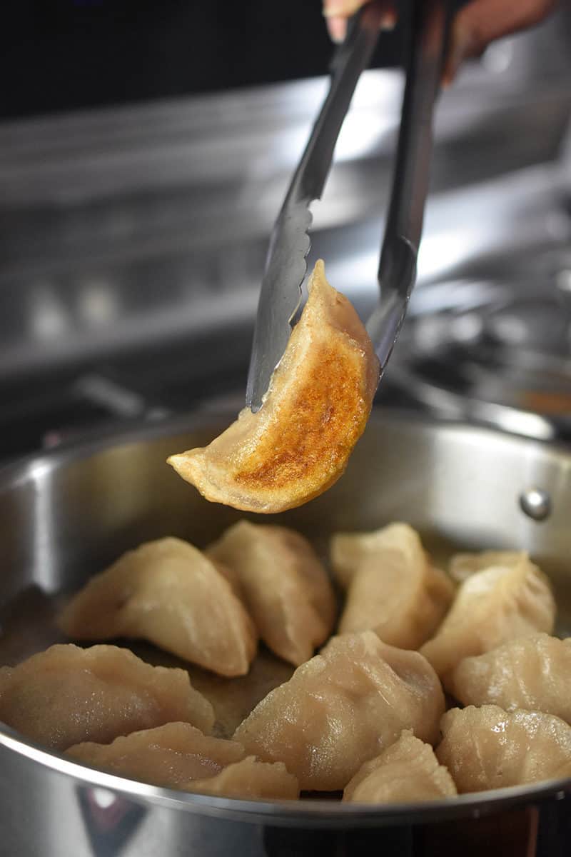 A pair of tongs is lifting up a cooked pot sticker, showing the crispy golden bottom.