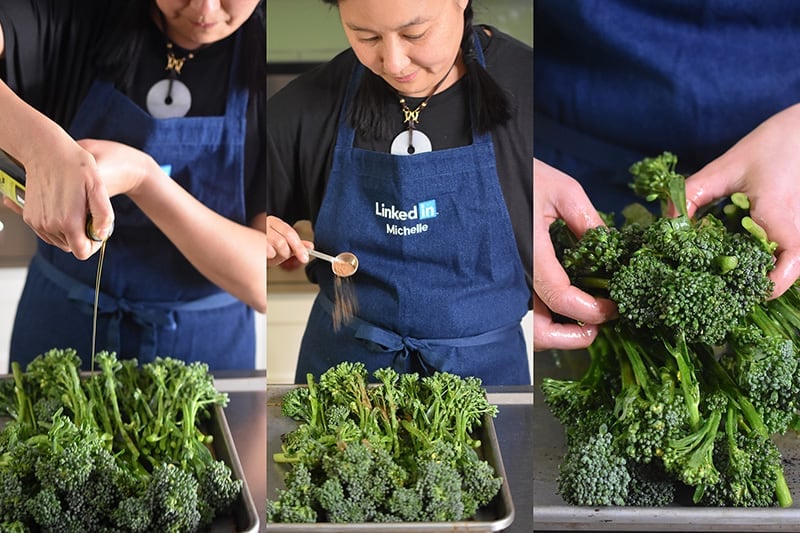A woman is tossing broccolini with olive oil and Magic Mushroom Powder on a rimmed baking sheet.