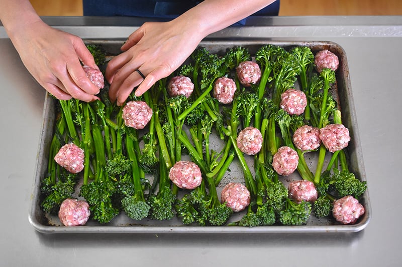 Two hands are shown adding meatballs to a rimmed baking sheet with broccolini.