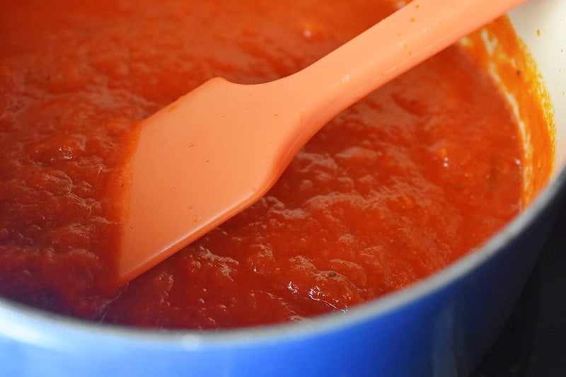 A close up of a silicone spatula stirring marinara sauce in a small Le Creuset saucepan.
