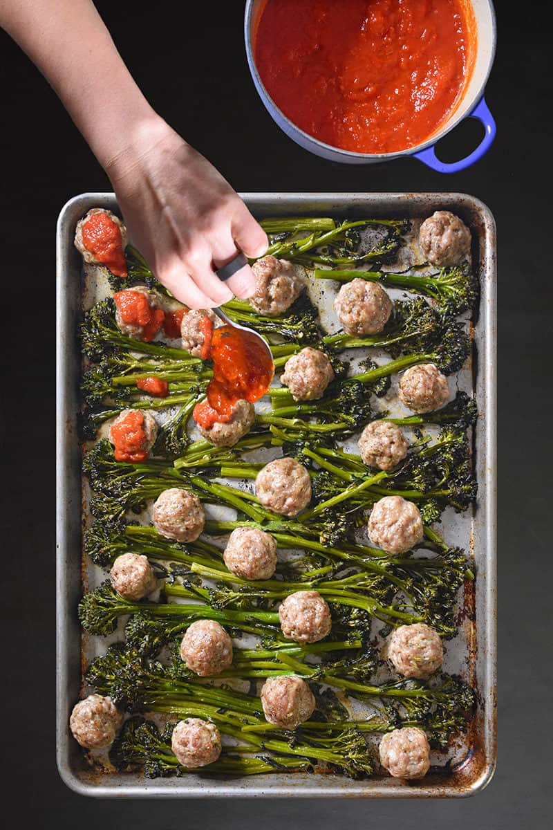Overhead shot of Sheet Pan Meatballs and Broccolini with someone spooning marinara sauce on top.