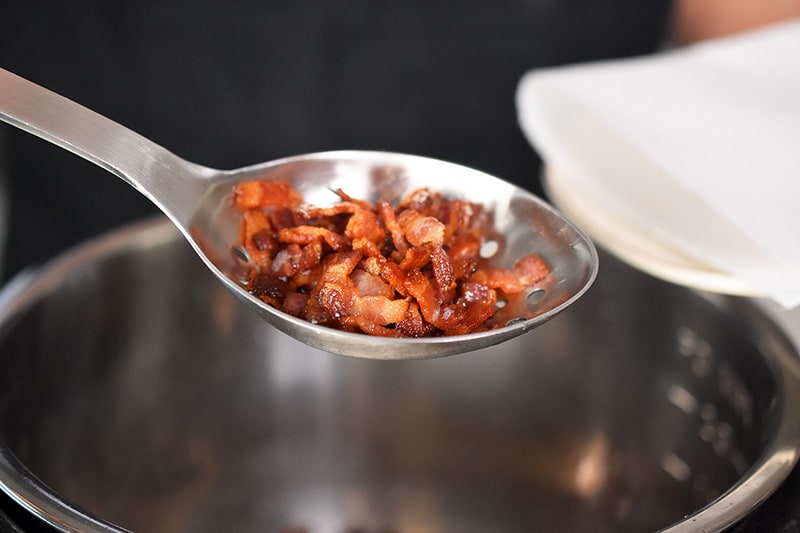 Crispy bacon bits are removed from the Instant Pot with a slotted spoon and transferred to a paper towel-lined plate.