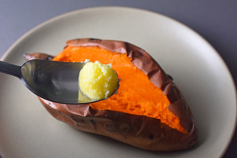 A baked sweet potato is split in half and a spoon with ghee is positioned on top.
